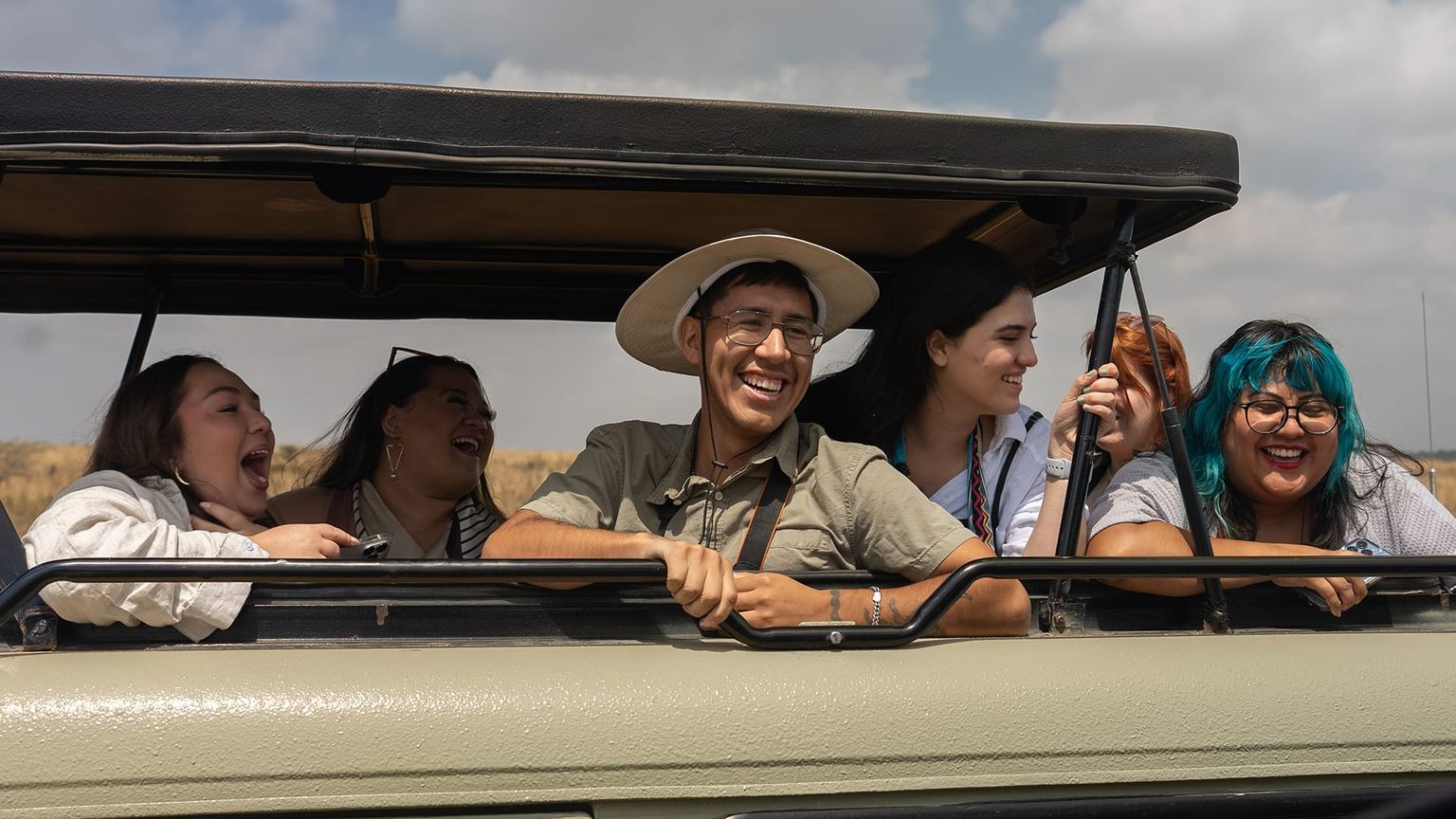 Five people smiling and laughing in an open-top safari vehicle under a cloudy sky. One wears a safari hat, and another has teal hair.