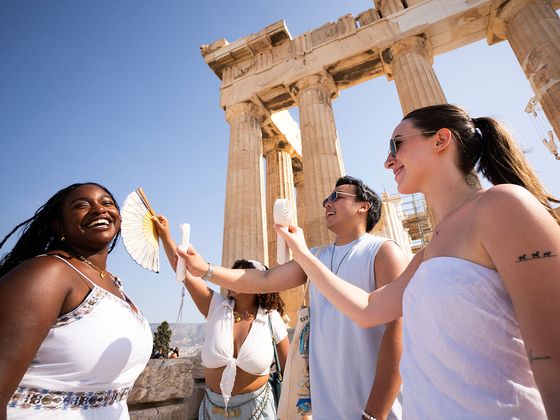 Four people enjoying a sunny day near the ancient Parthenon, smiling as they use fans to cool off.