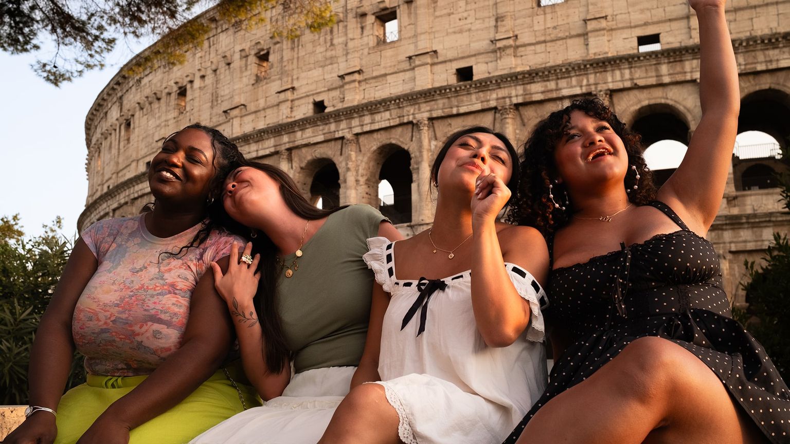 Four women sit and laugh together in front of the Colosseum, with the sun casting a warm glow on the scene.