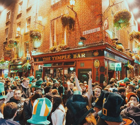 A bustling crowd celebrates outside a lively pub at night, adorned with Irish flags and decorations, under warm streetlights.