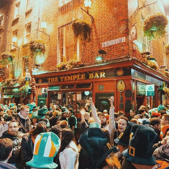 A bustling crowd celebrates outside a lively pub at night, adorned with Irish flags and decorations, under warm streetlights.