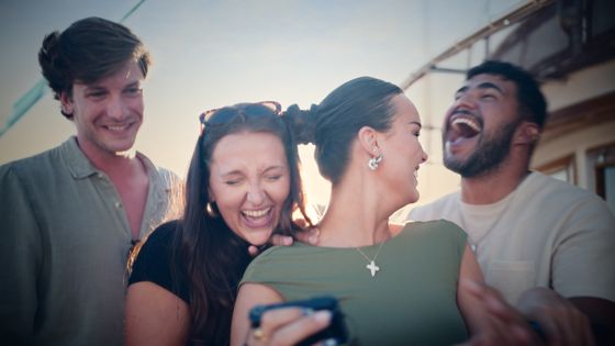 A group of four friends laughing together on a boat under a clear sky, with two women in the center and two men on either side.