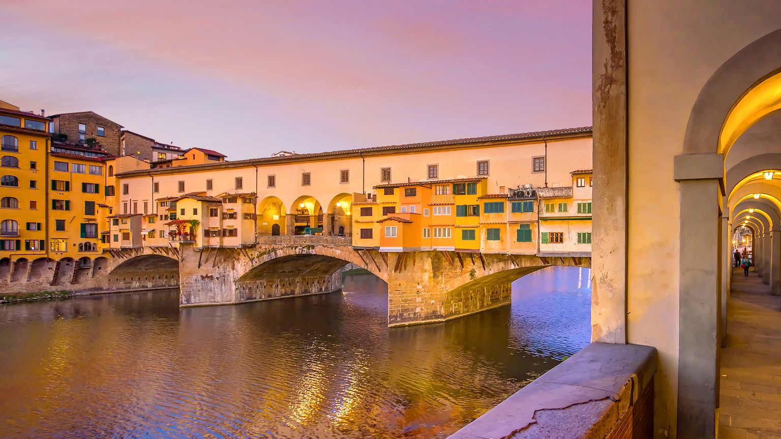 Sunset view of Ponte Vecchio, an ancient bridge with colorful buildings, spanning the Arno River in Florence, Italy, with arched walkway in foreground.