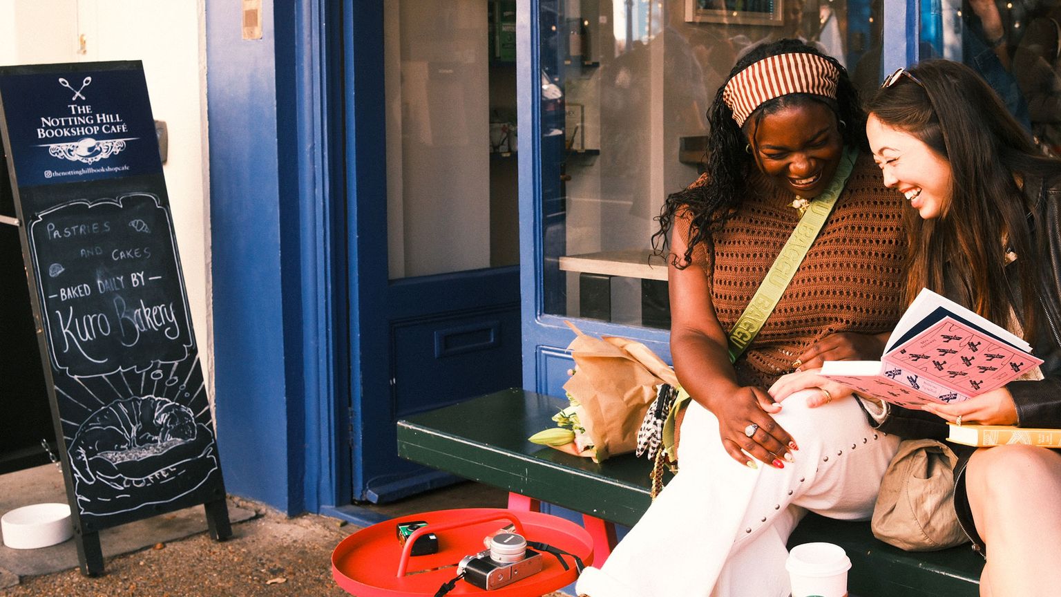 Two friends laughing on a bench outside a blue bookshop café, sharing a pink book with coffee and flowers beside them.