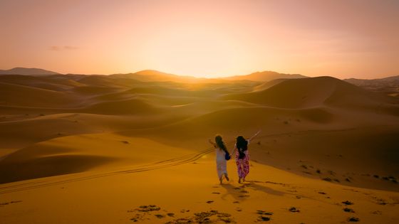 Two people walking hand in hand through golden desert dunes at sunset, leaving footprints in the sand.