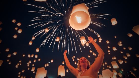A person joyfully raises arms among floating lanterns, with a dazzling firework display lighting up the night sky.