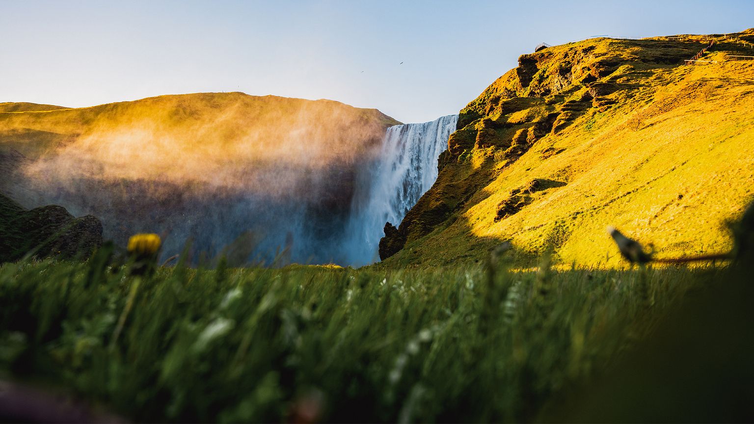 A majestic waterfall cascades down a rocky cliff into lush green grass, with sunlight casting warm, golden hues over the scene.