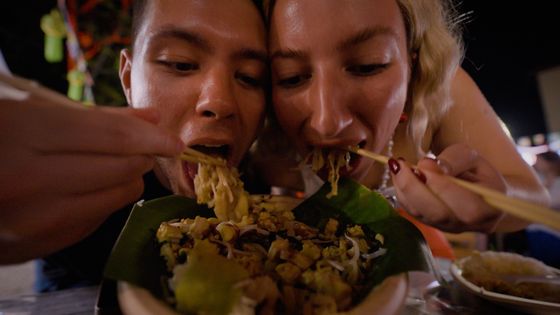 Two people eating noodles from a shared bowl with chopsticks, surrounded by a lively, colorful environment.