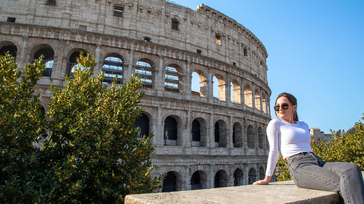 A woman in sunglasses sits on a ledge in front of the Colosseum in Rome, with clear blue skies and green foliage nearby.
