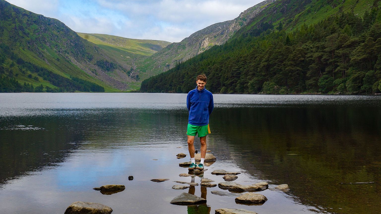 A person stands on stepping stones in a calm lake, surrounded by lush green hills and forests under a partly cloudy sky.