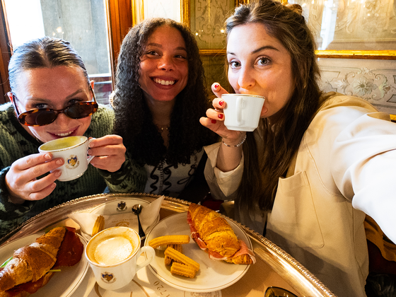 Three women enjoying coffee and pastries at a café, smiling and sitting at a table with ornate wall decorations.