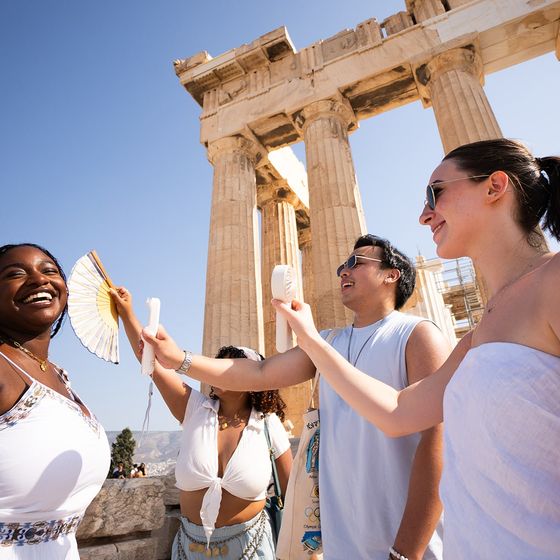 Four friends smile and enjoy the sunny day in front of ancient ruins, fanning themselves to stay cool.