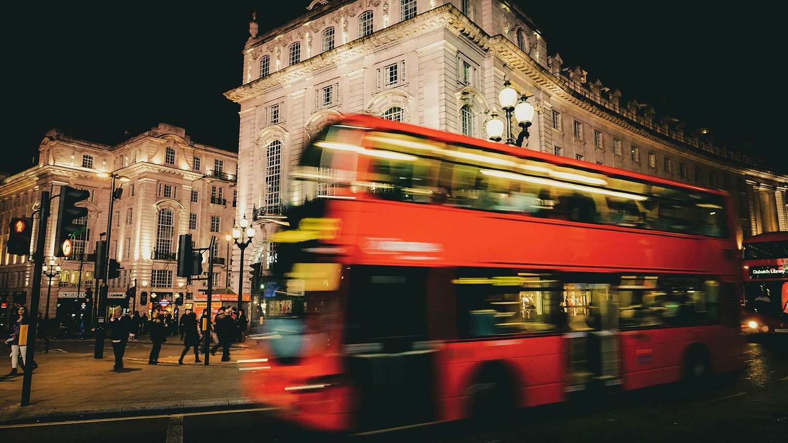 A blurred red double-decker bus passes by illuminated historic buildings at night in a bustling city scene.