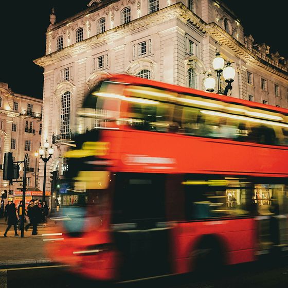 A blurred red double-decker bus passes by illuminated historic buildings at night in a bustling city scene.