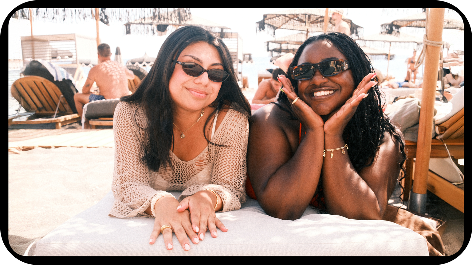 Two women wearing sunglasses, smiling at the camera, relaxing on a beach lounge chair. Beach umbrellas and other people in the background.