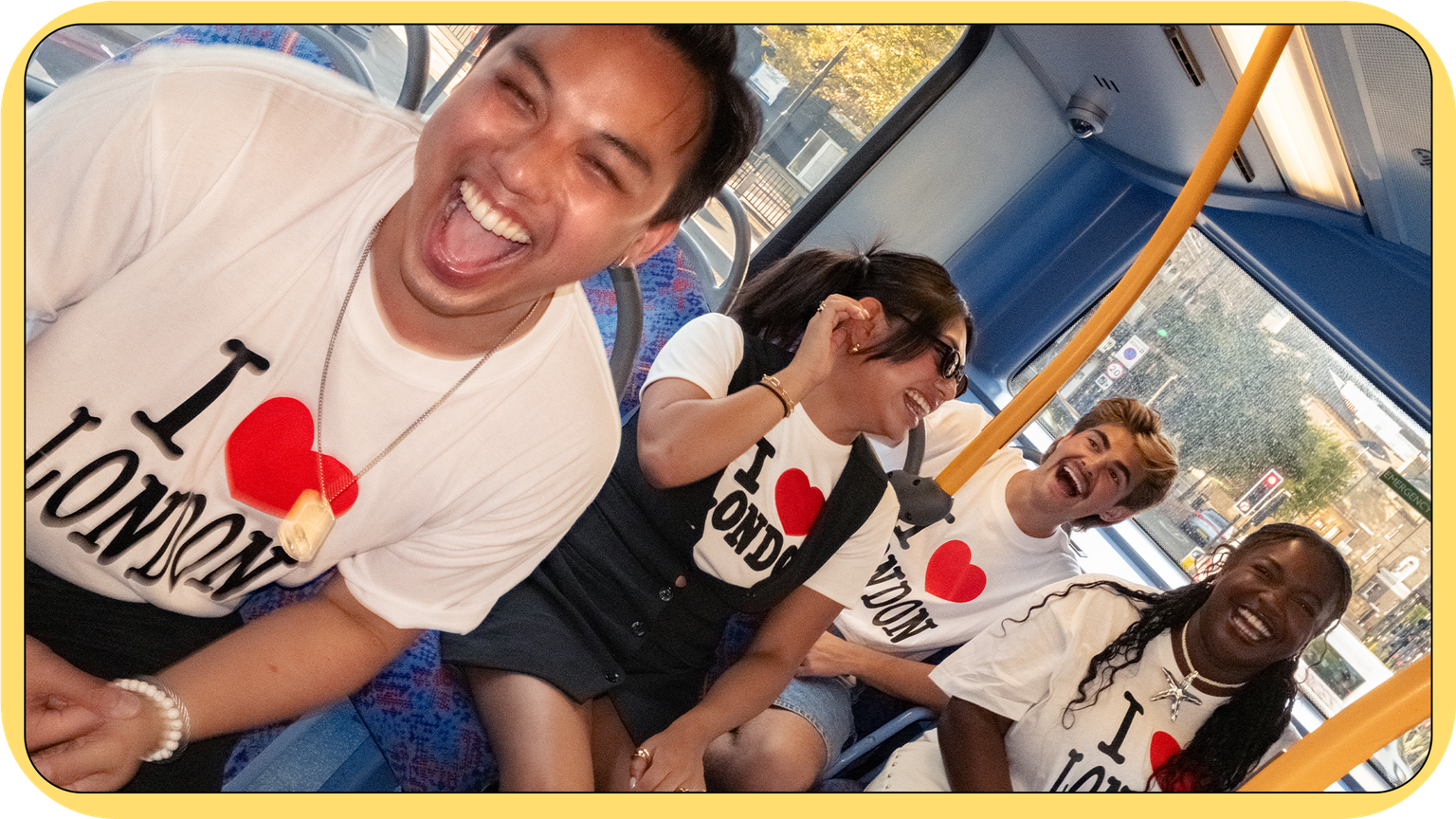 Four people wearing "I ♥ London" shirts are laughing and enjoying themselves on a bus with blue patterned seats and yellow poles.