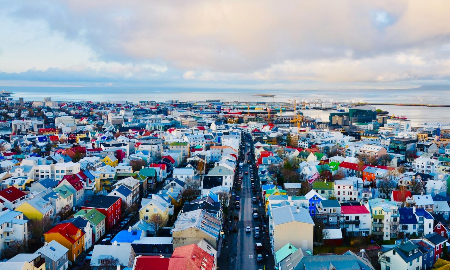 Aerial view of a coastal city with colorful houses, a busy street, and a harbor in the background under a partly cloudy sky.