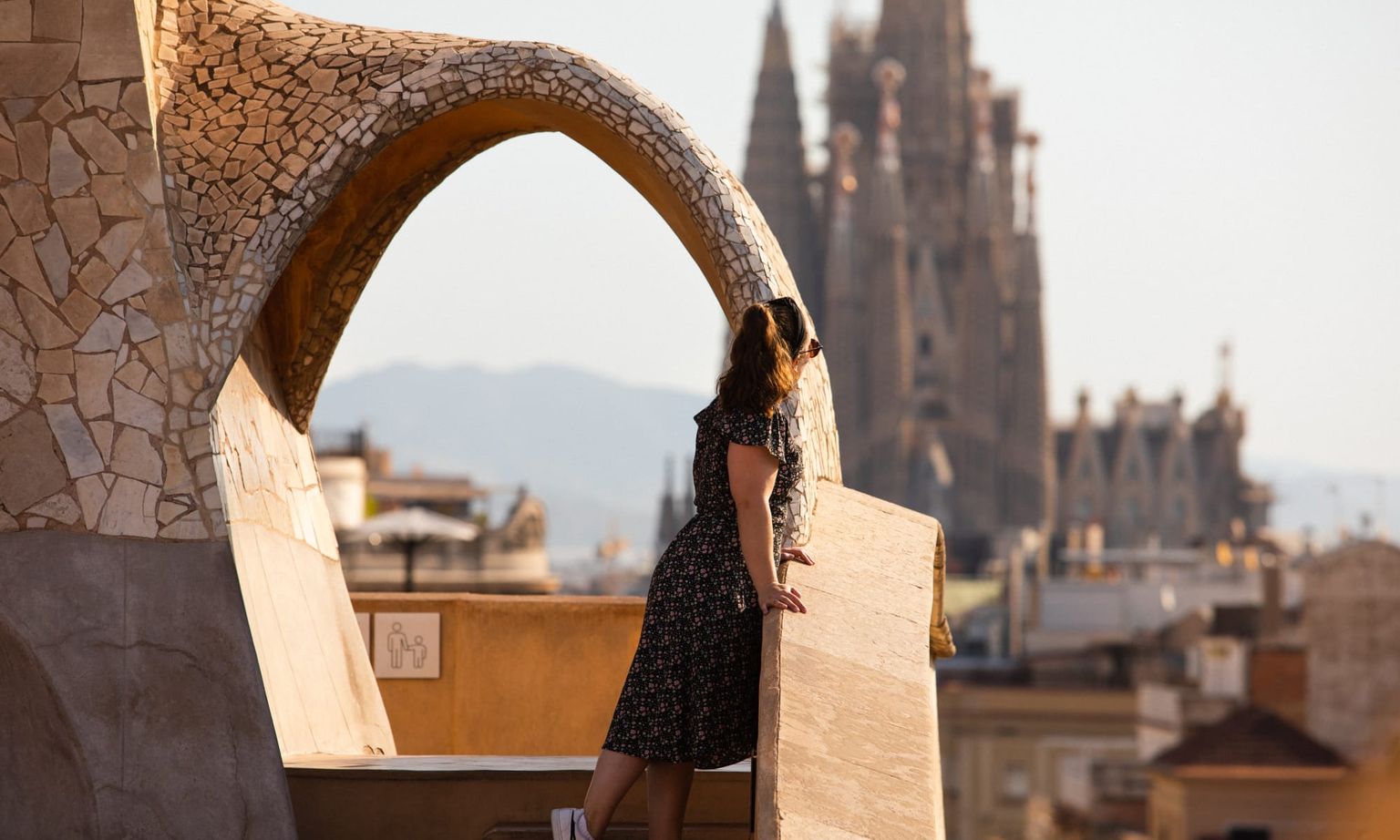A woman standing in the sun on a stone staircase looking toward the Sagrada Familia in Barcelona, Spain