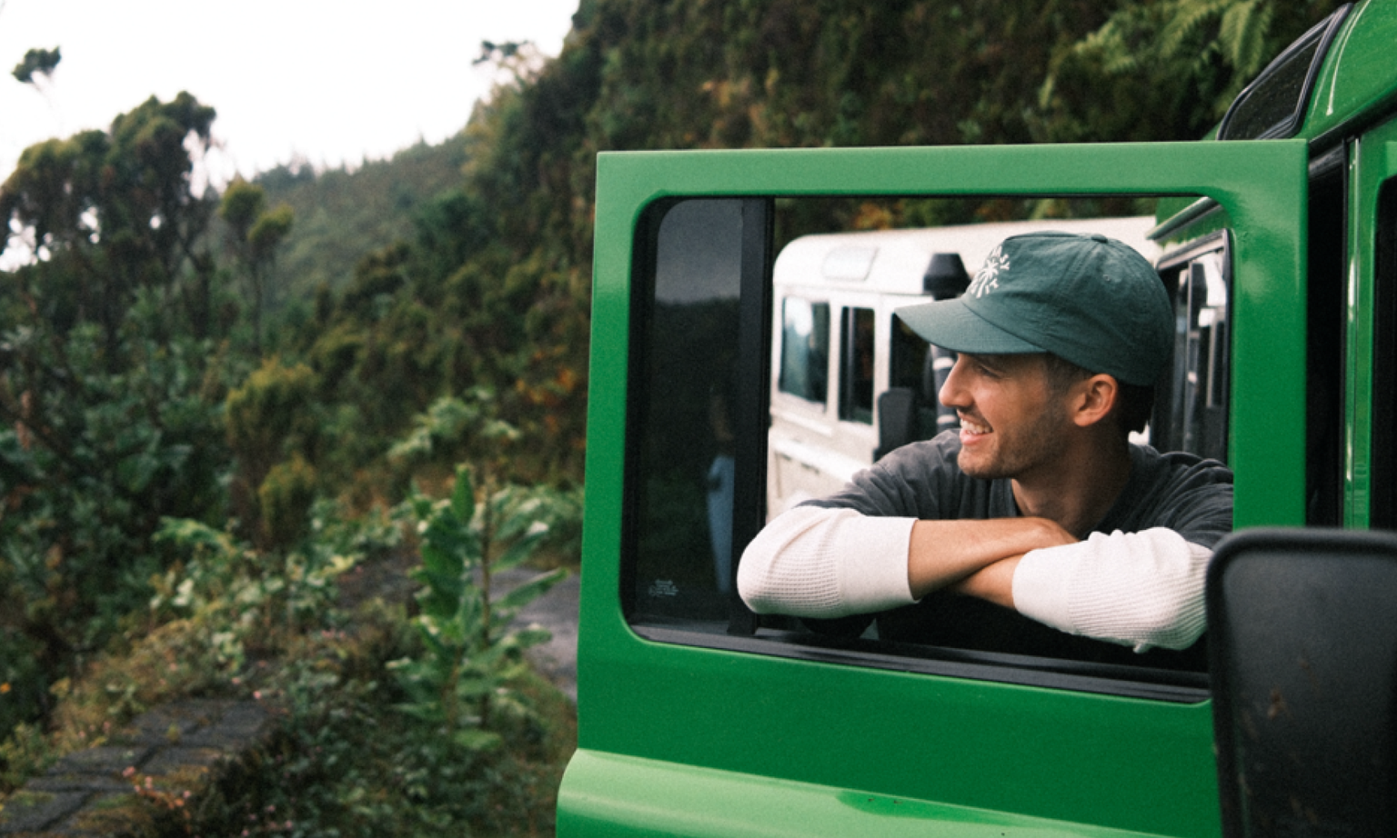 A man standing in the window of a green jeep surrounded by lush jungle and smiling off into to the distance