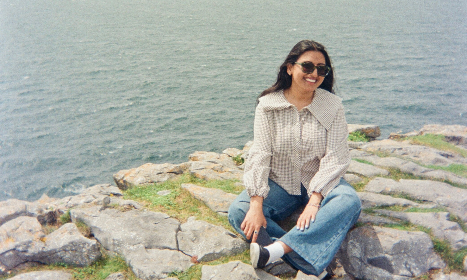 Woman in sunglasses sitting on rocky shore, wearing a checkered blouse and jeans, smiles with the ocean in the background.