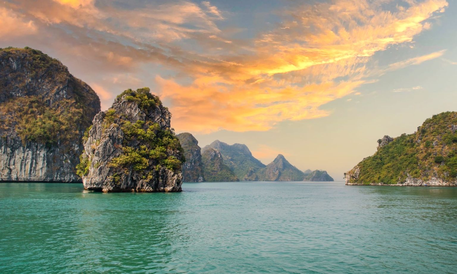 Scenic view of rocky islands covered with greenery in a calm turquoise sea under a vibrant orange and pink sunset sky.
