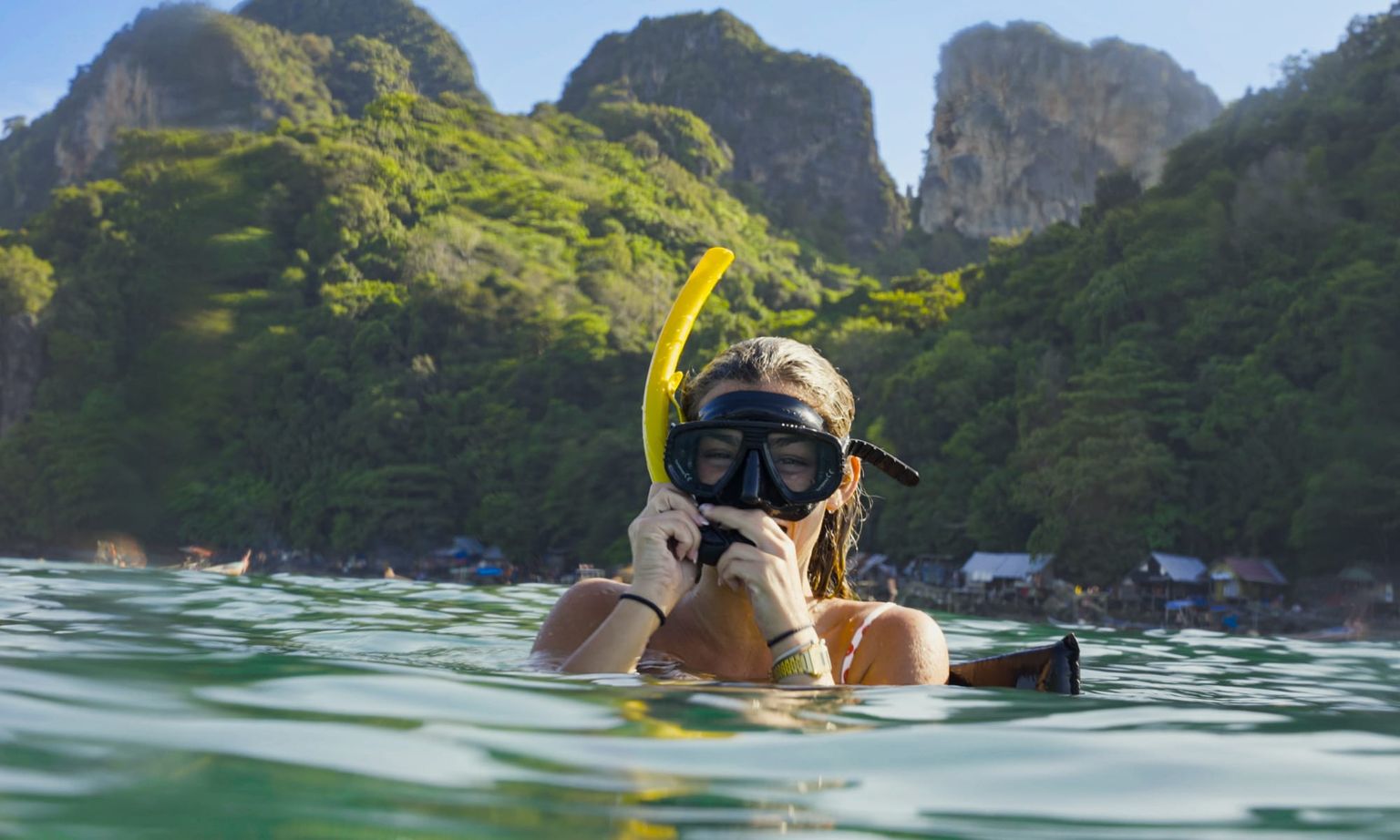 A person snorkeling in clear water with lush green mountains and a distant village visible in the background.