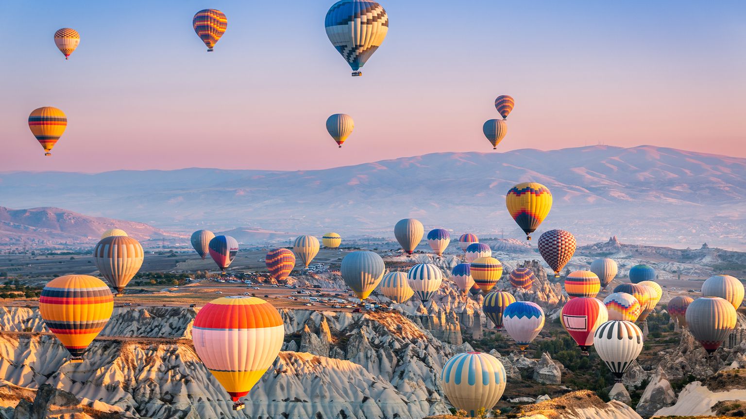 Many colorful hot air balloons floating over the rock formations of Cappadocia, Turkey with mountains in the distance