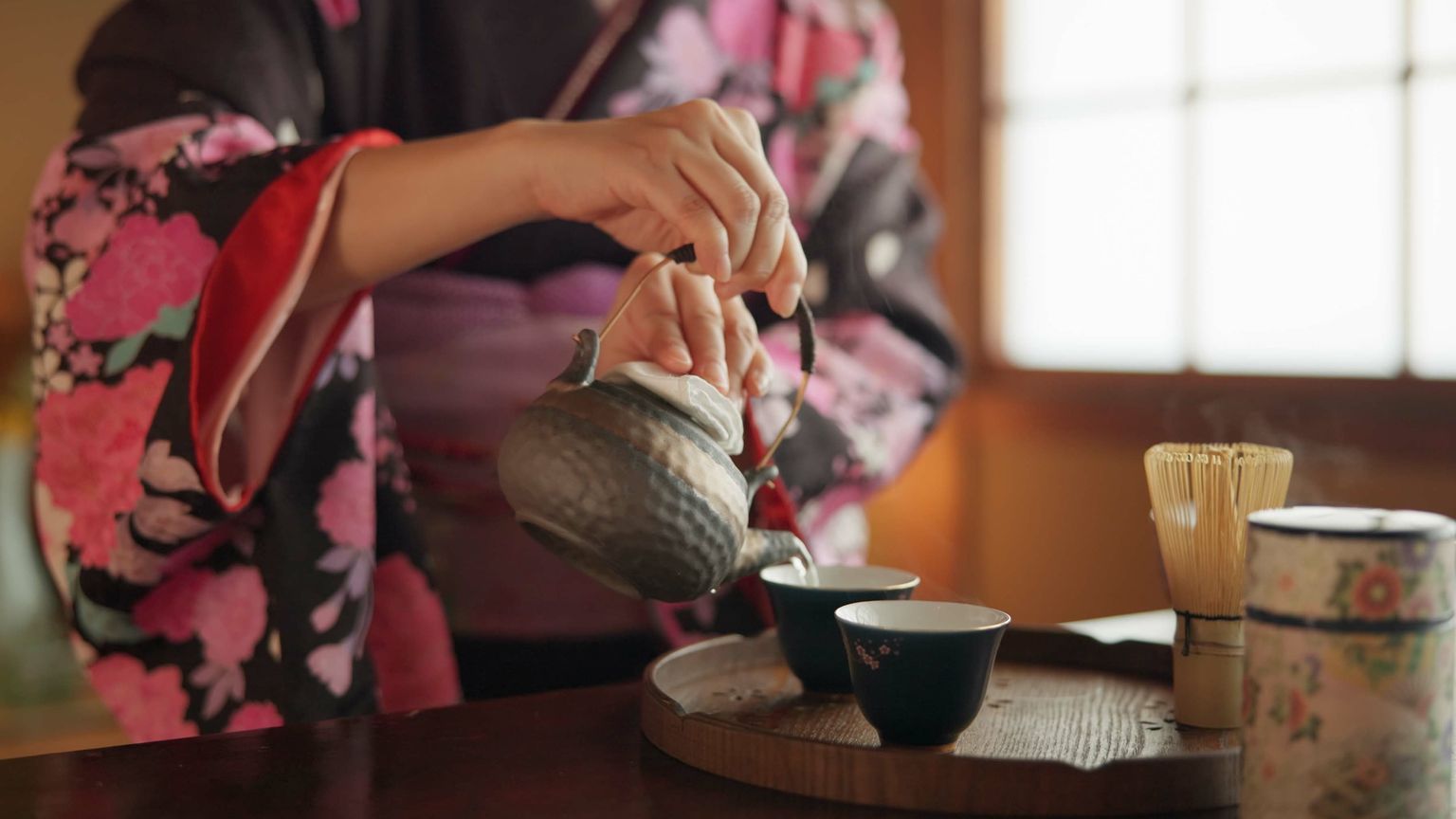 A person in a floral kimono pours tea from a traditional teapot into cups on a tray, with a bamboo whisk and tea canister nearby.