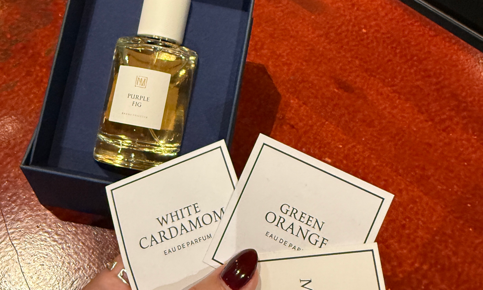 A bottle of perfume in a box and a hand holding paper cards printed with different perfume scents