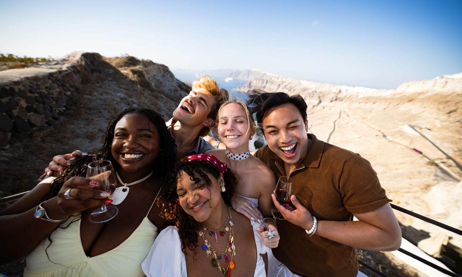 Group of five people smiling and holding drinks, posing against a rocky, sunny landscape with clear blue skies in the background.