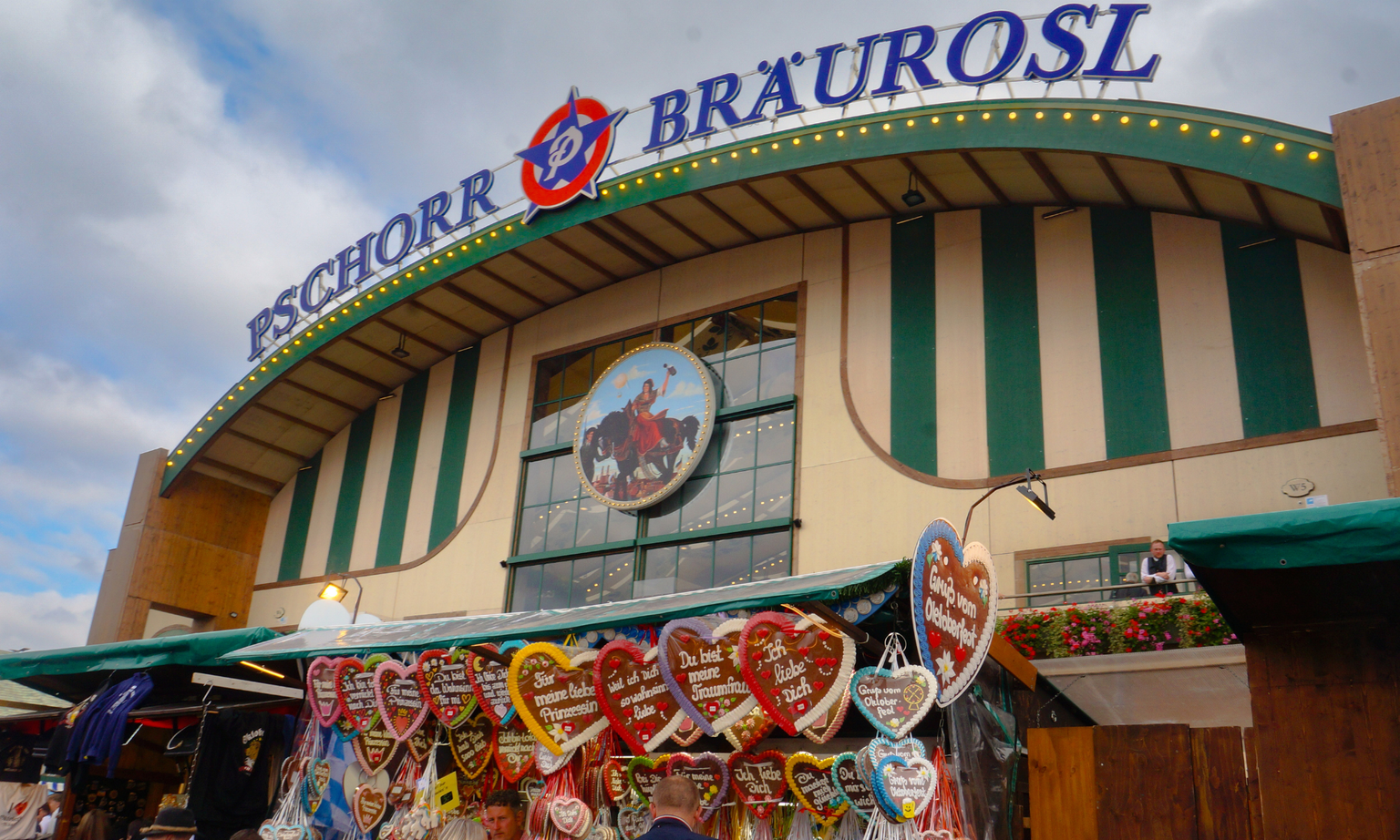 Oktoberfest tent with "Pschorr Bräurosl" sign, striped facade, and a stall displaying heart-shaped gingerbread cookies.