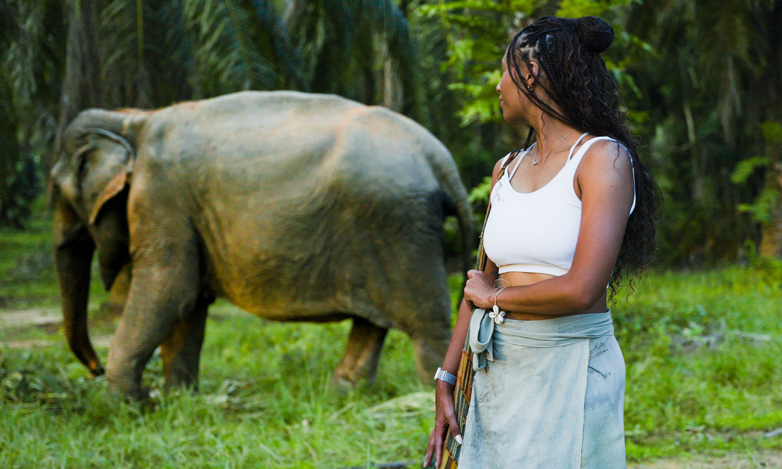 A woman looking away from the camera toward an elephant in the background at an ethical Thai sanctuary