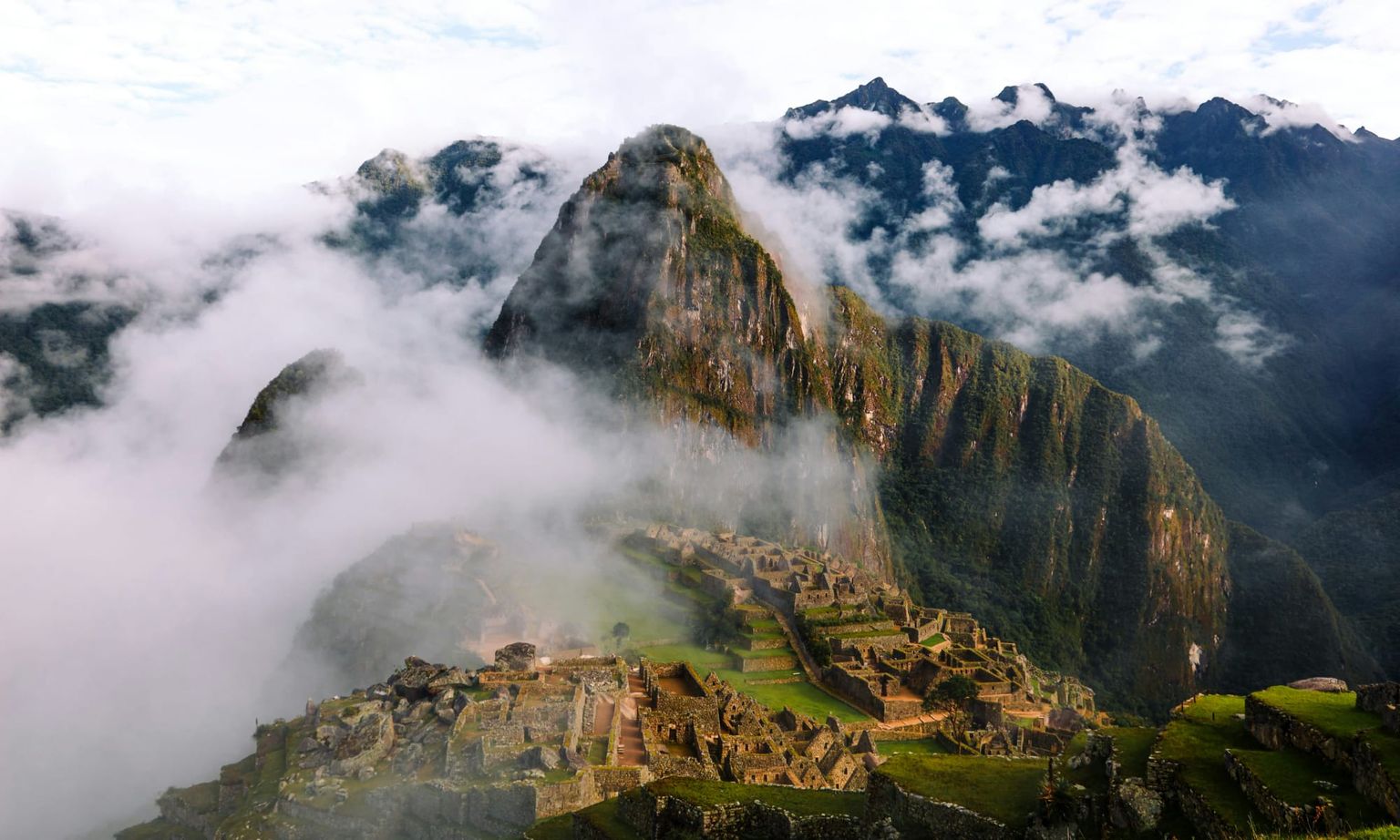 Machu Picchu with misty clouds surrounding the ancient ruins, set against the backdrop of towering mountains under a partly cloudy sky.