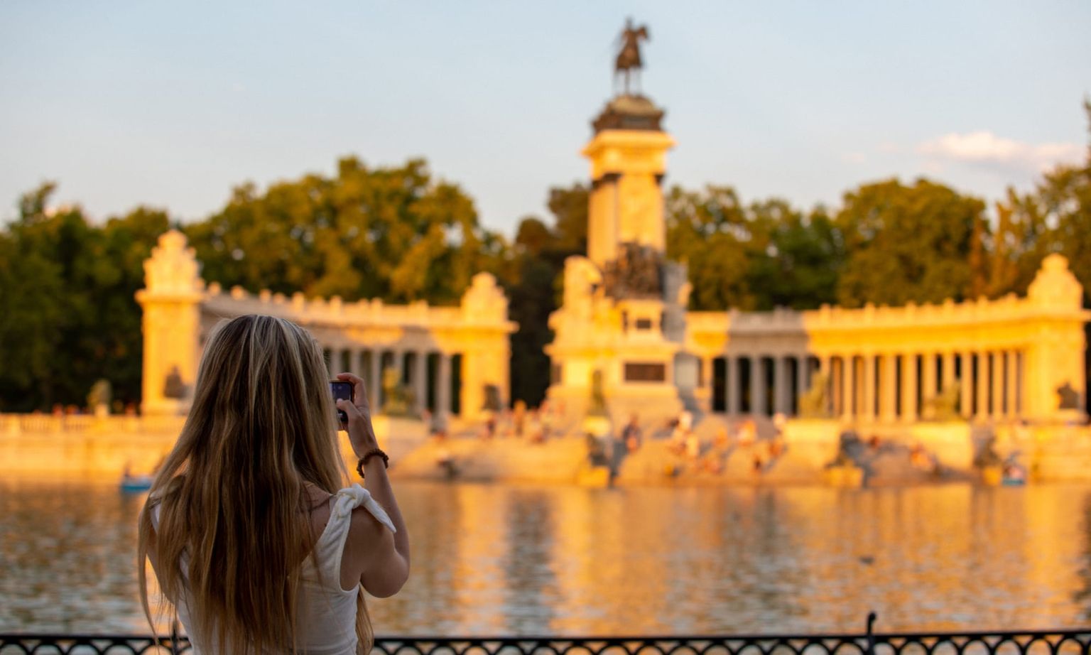 Woman photographing a monument across a sunlit lake, with trees and people in the background at sunset.