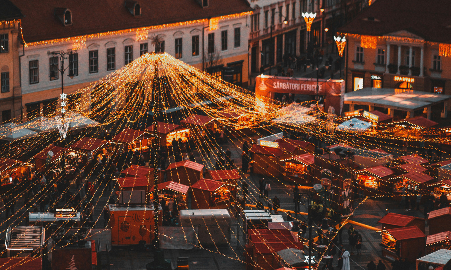 Festive market square adorned with string lights, red stalls, and illuminated buildings, creating a warm, inviting holiday atmosphere.