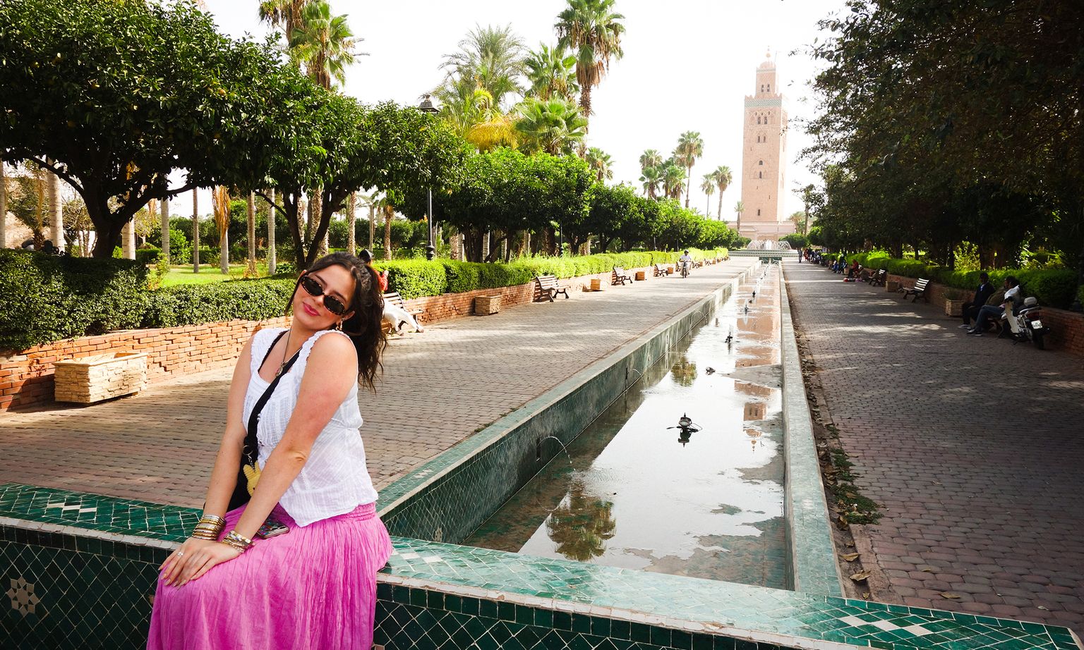 Woman in a white top and pink skirt poses by a reflecting pool in a park with palm trees and a tall tower in the background.