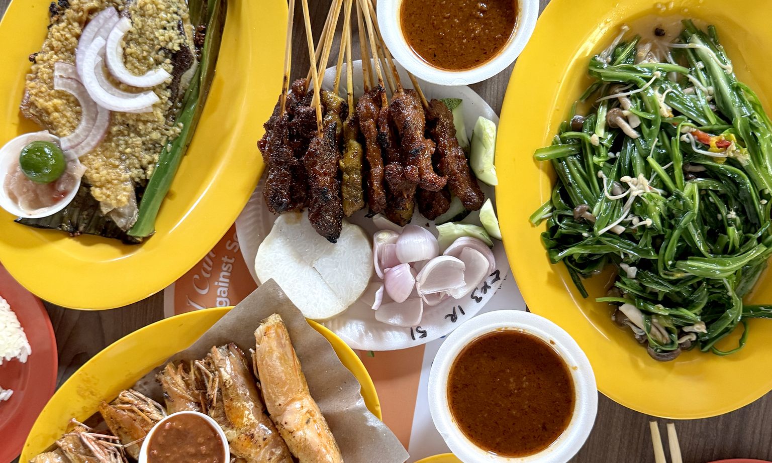 A variety of Malaysian dishes on yellow plates, including grilled skewers, prawns, vegetables, sauces, and garnishes.
