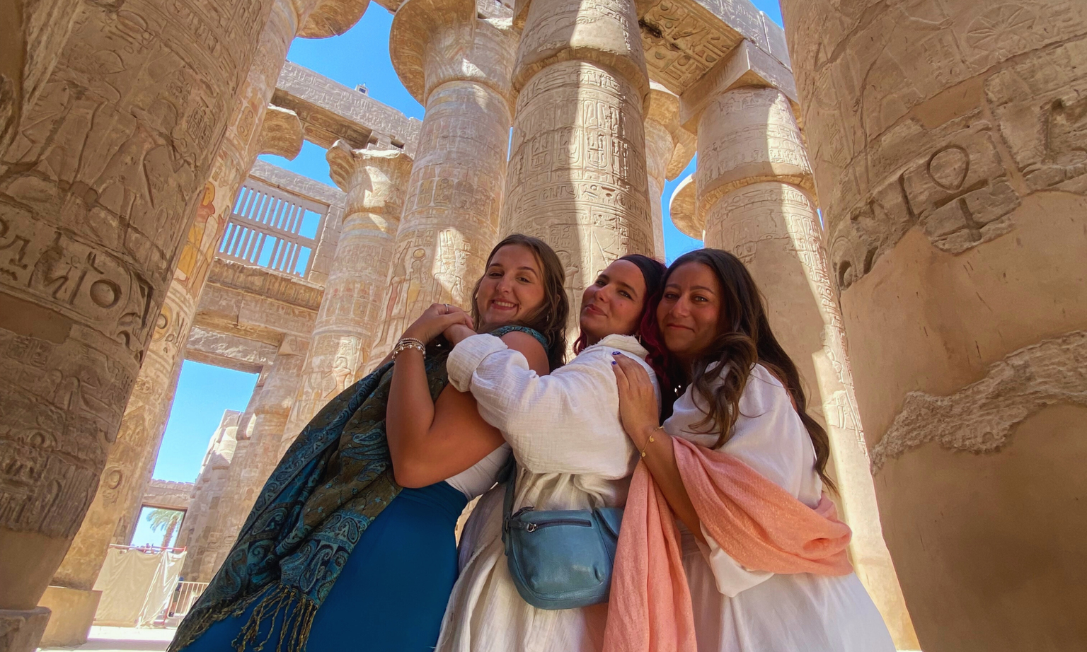 Three women embracing and smiling surrounded by hieroglyph-covered columns in Egypt