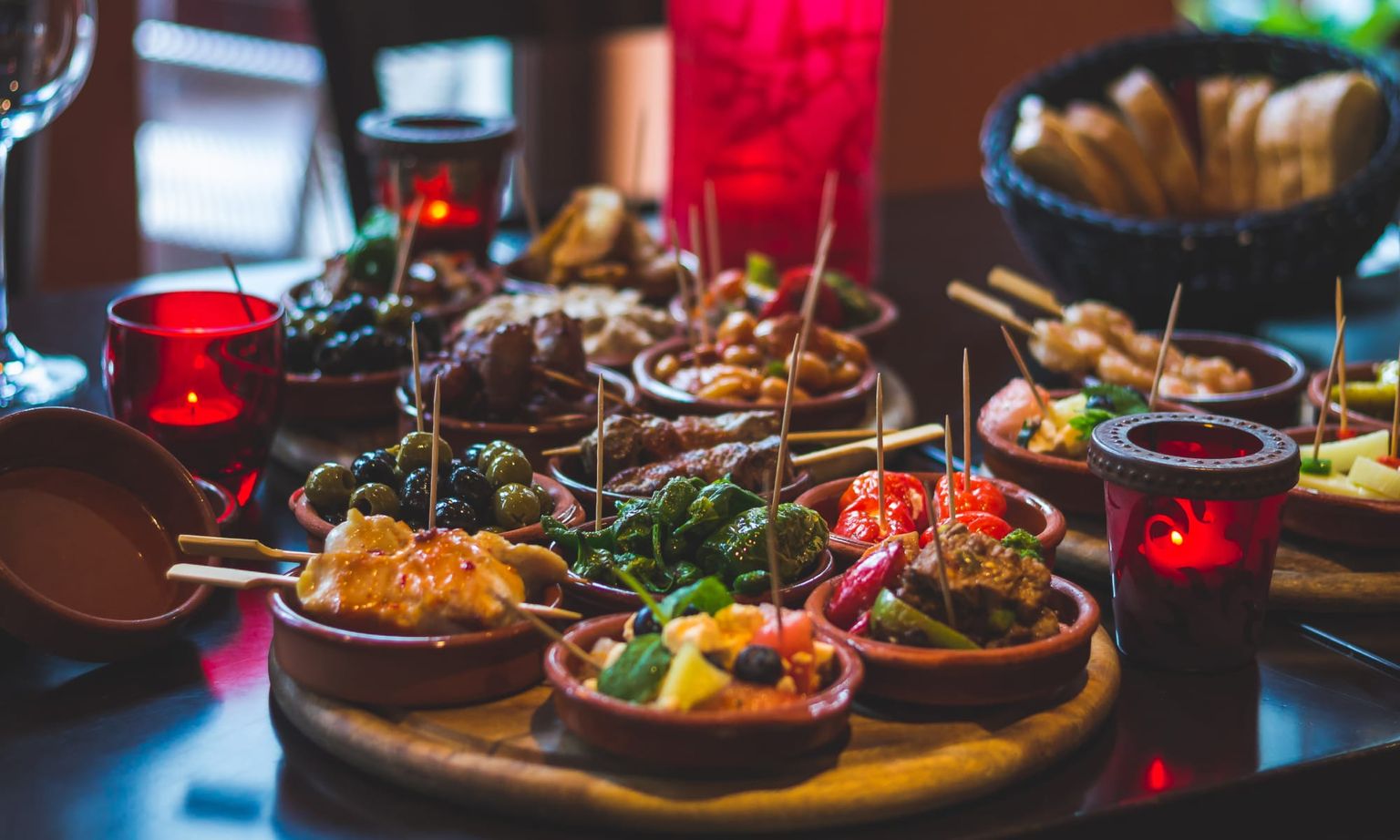 A table filled with various tapas dishes, including olives, meats, and vegetables, surrounded by red candles and bread in a basket.