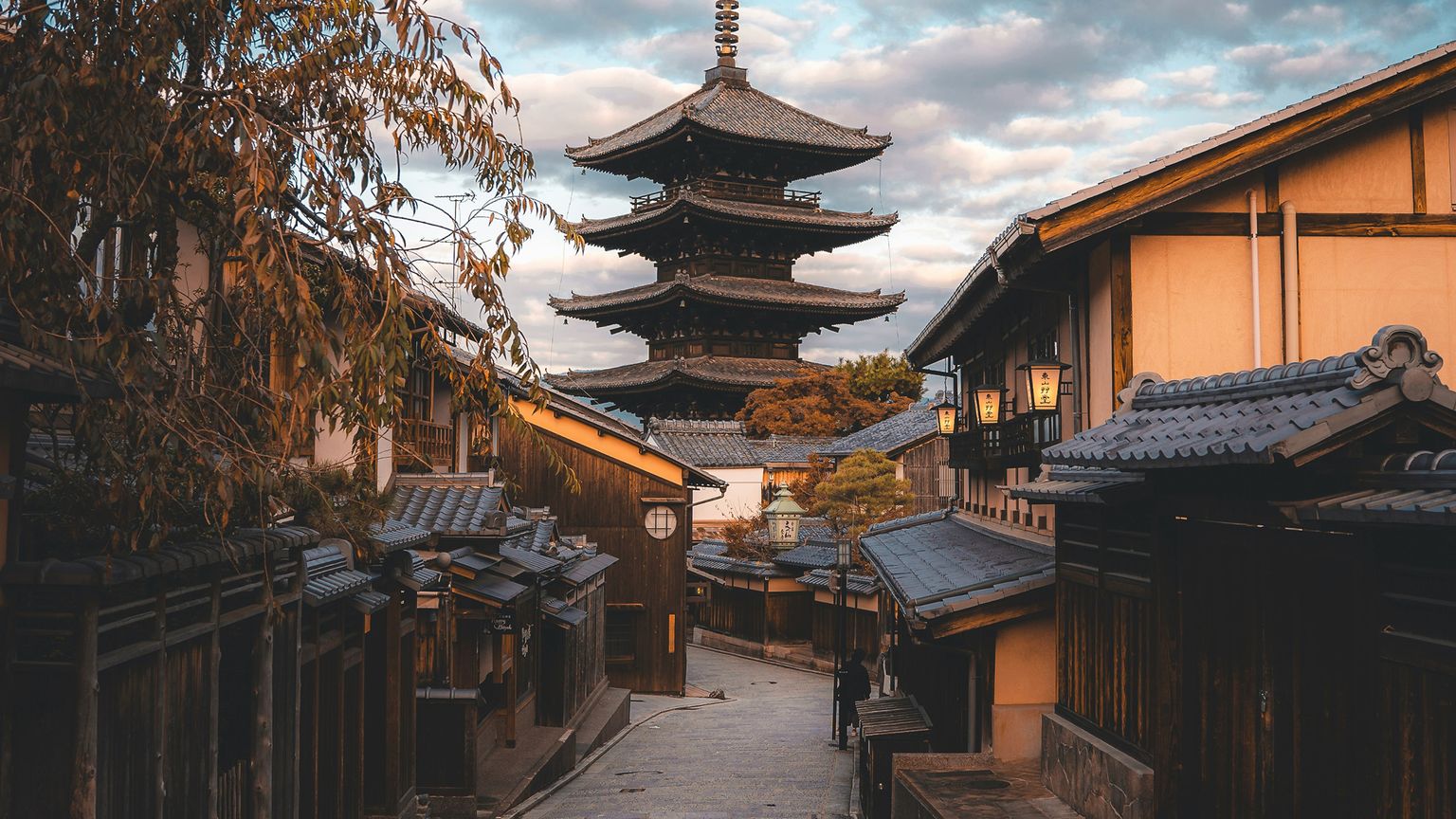 A narrow street with traditional wooden buildings leads to a tall pagoda under a partly cloudy sky at sunset.