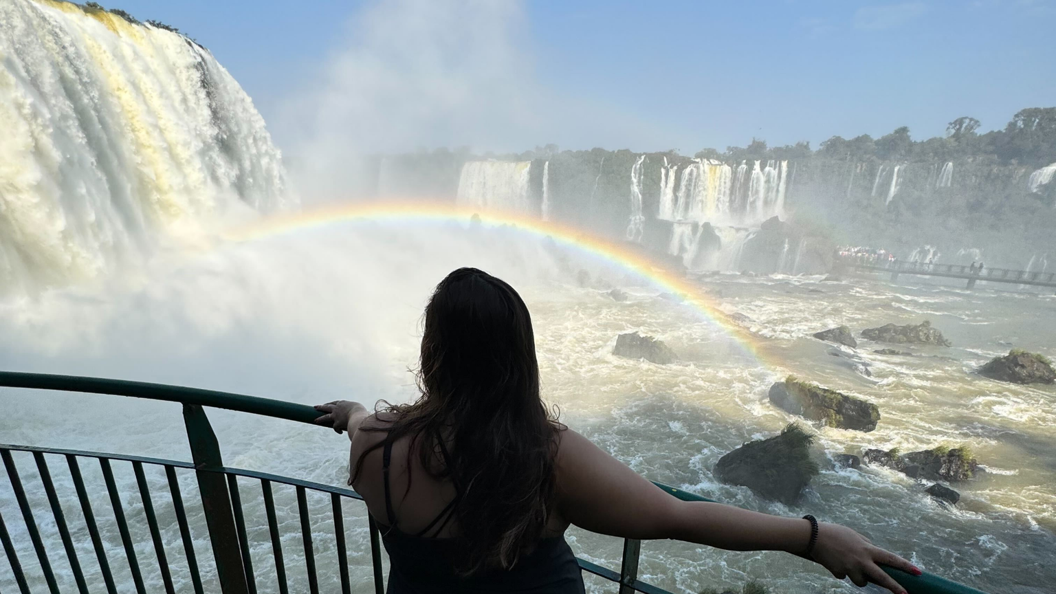 A woman facing away from the camera and looking toward a huge waterfall and rainbow