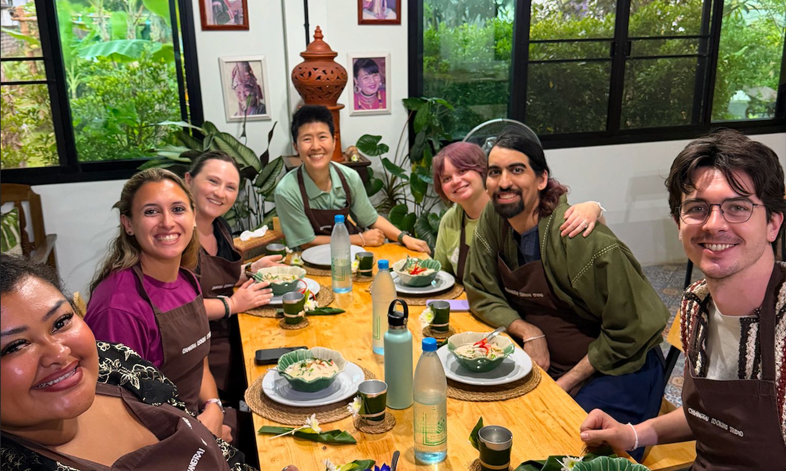 A group of people at a table, smiling with bowls of food. They are in a cozy room with plants and photos on the walls.