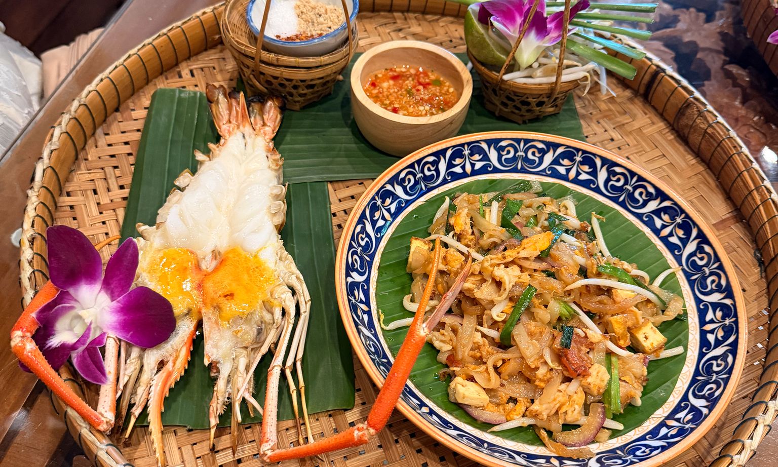 A woven tray with grilled prawns, a plate of pad Thai, dipping sauce, and a decorative orchid on banana leaves.