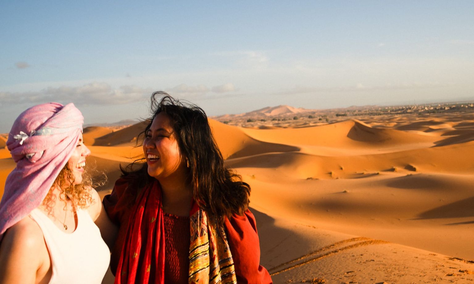 Two smiling women stand in a sandy desert under a clear sky, one wearing a pink headscarf and the other in a red outfit with a scarf.