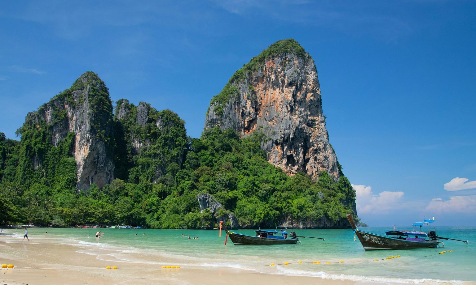A scenic beach with turquoise waters, longtail boats, and lush green cliffs under a clear blue sky.