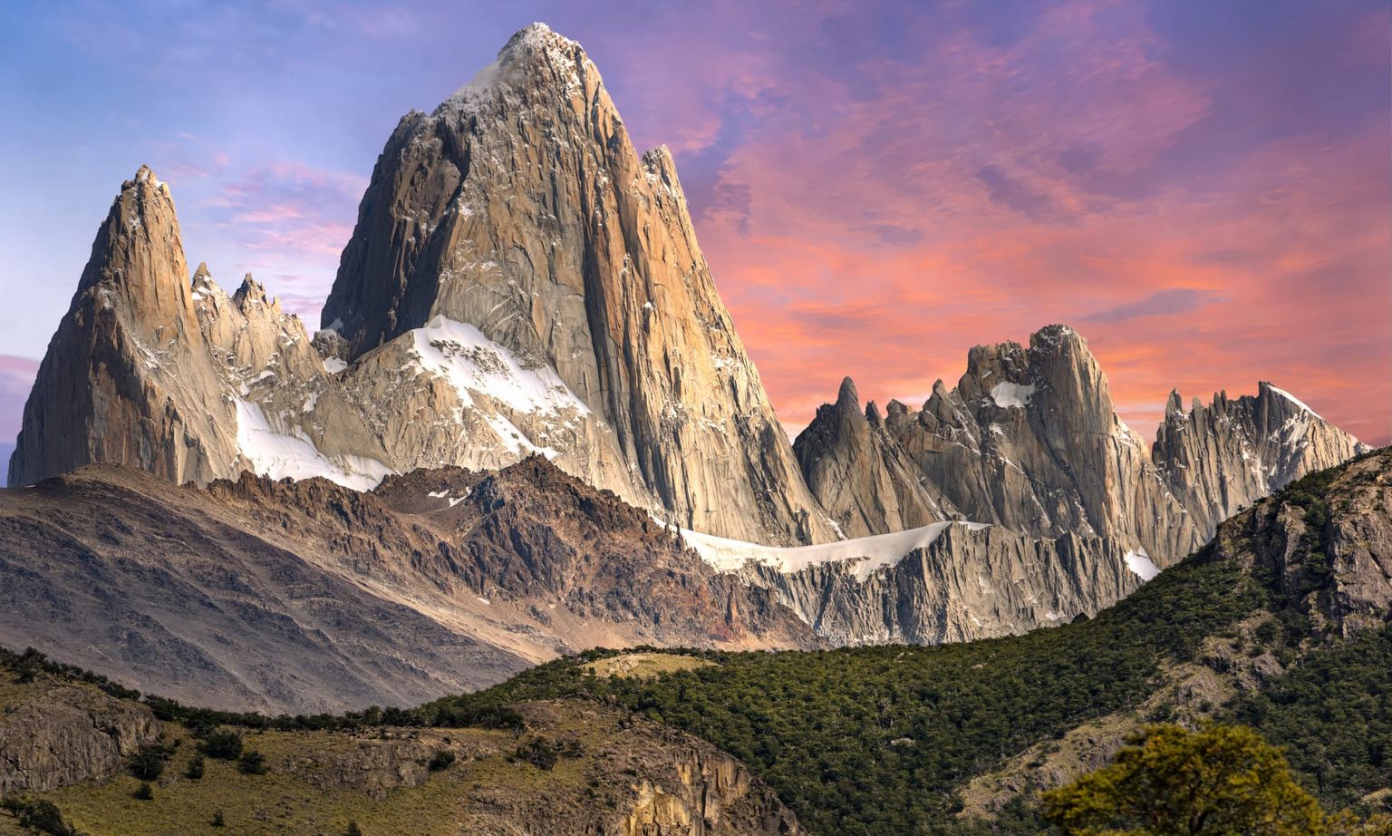 Sunset over Mount Fitz Roy in Patagonia, with rugged peaks glowing against a pink and orange sky, and forested slopes below.