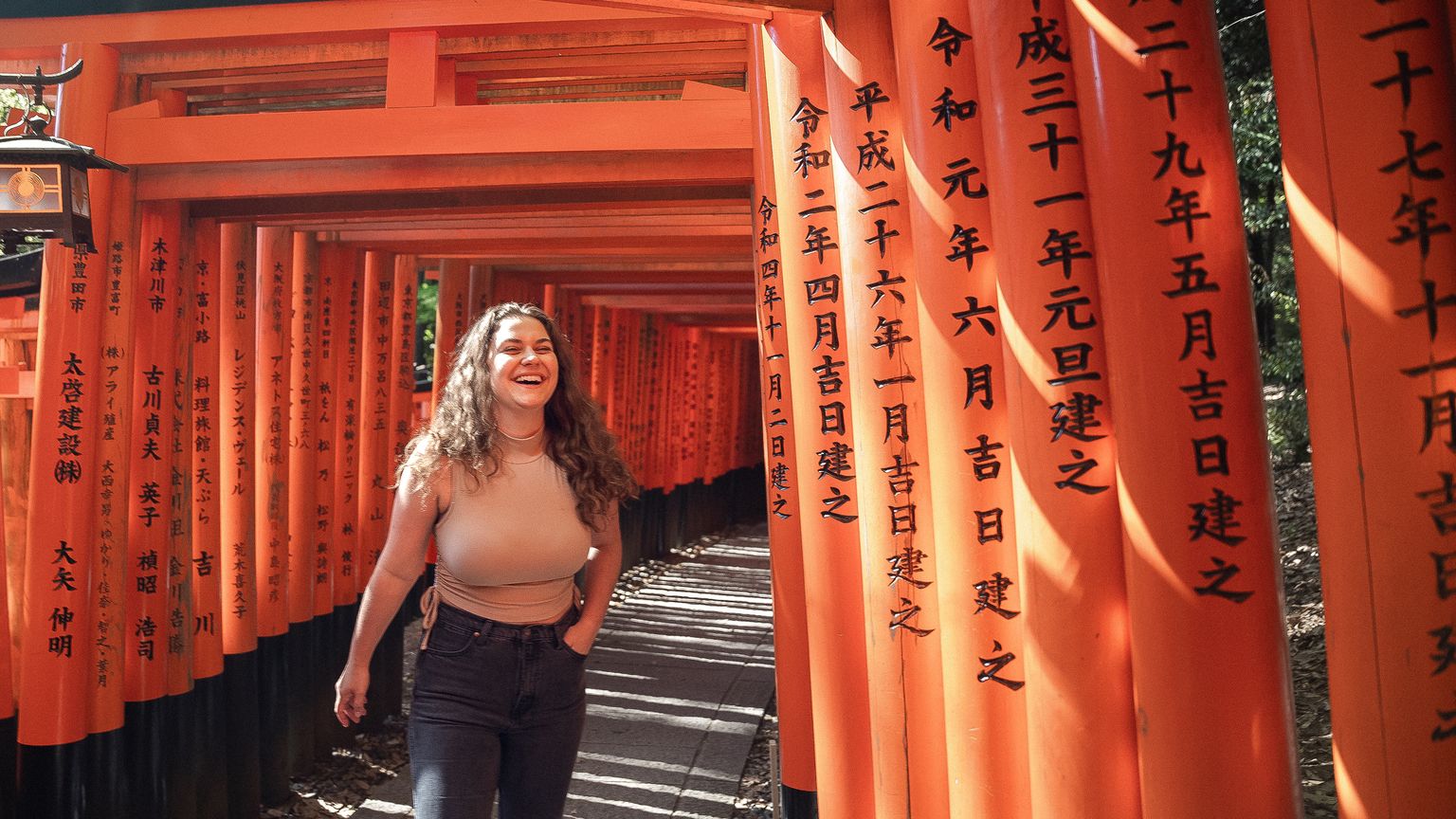 A person smiling while walking through orange Torii gates carved with Japanese characters at Kyoto's Fushimi Inari Taisha shrine