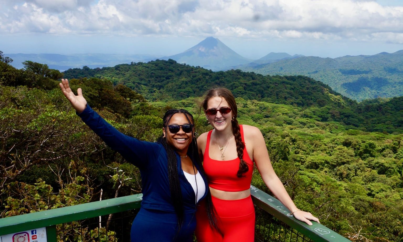 Two women smiling and posing on a balcony overlooking lush green hills, with a distant volcanic mountain under a cloudy sky.