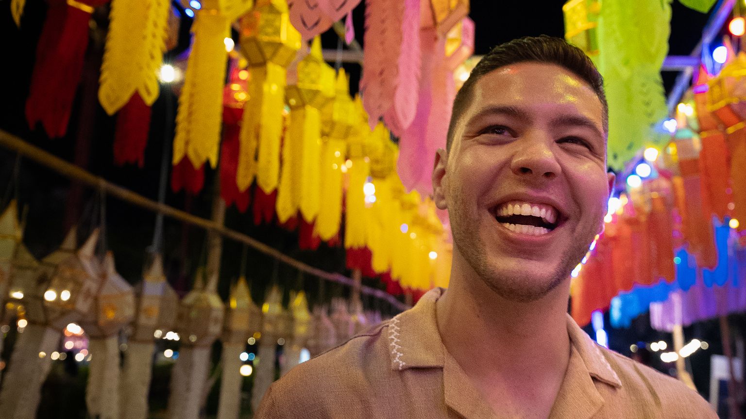 A person smiling widely against a backdrop of colorful hanging lanterns and lights at night.