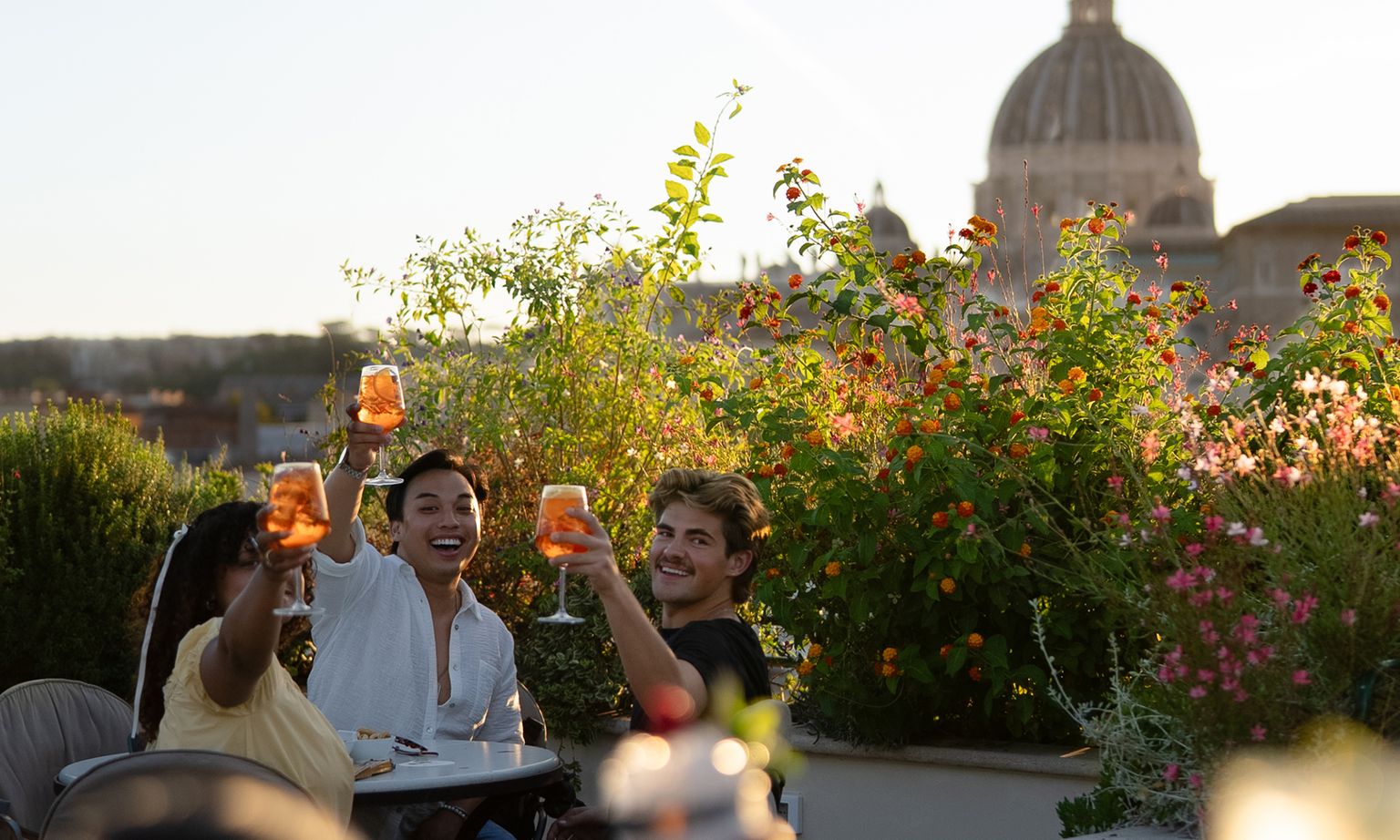 Three friends toast with drinks on a rooftop terrace, surrounded by lush plants, with a large domed building in the background.